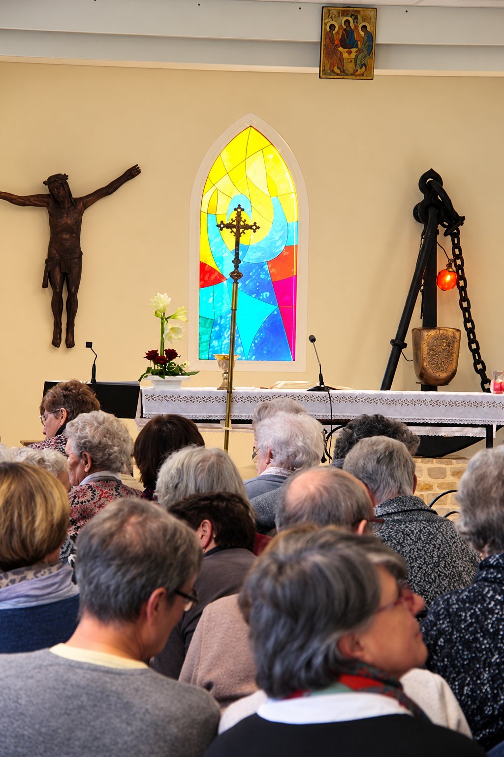 Des fidèles vus de dos assistent à une célébration dans une chapelle, face à un autel drapé de blanc, un vitrail coloré, un crucifix et une icône au mur.