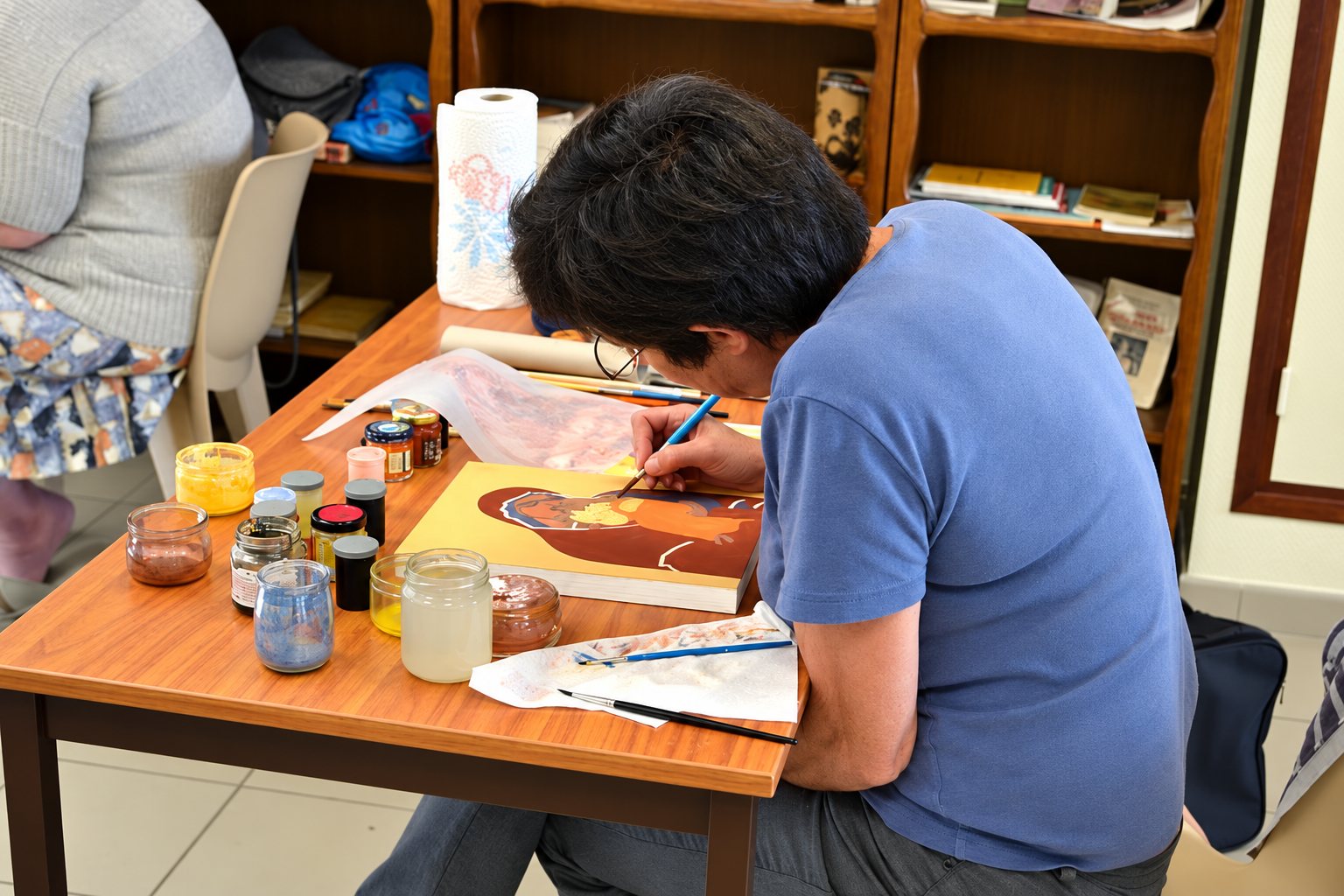 Une personne en t-shirt bleu peint une icône sur un panneau de bois à une table couverte de pots de pigments et de pinceaux, dans une salle avec des étagères de livres.