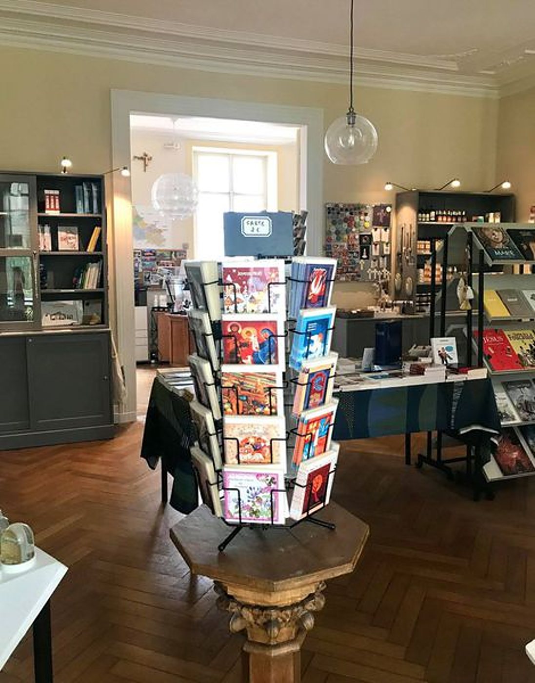 Intérieur lumineux d'une boutique avec un présentoir tournant de cartes postales sur un pied en bois, des étagères de livres et un parquet en chevrons.