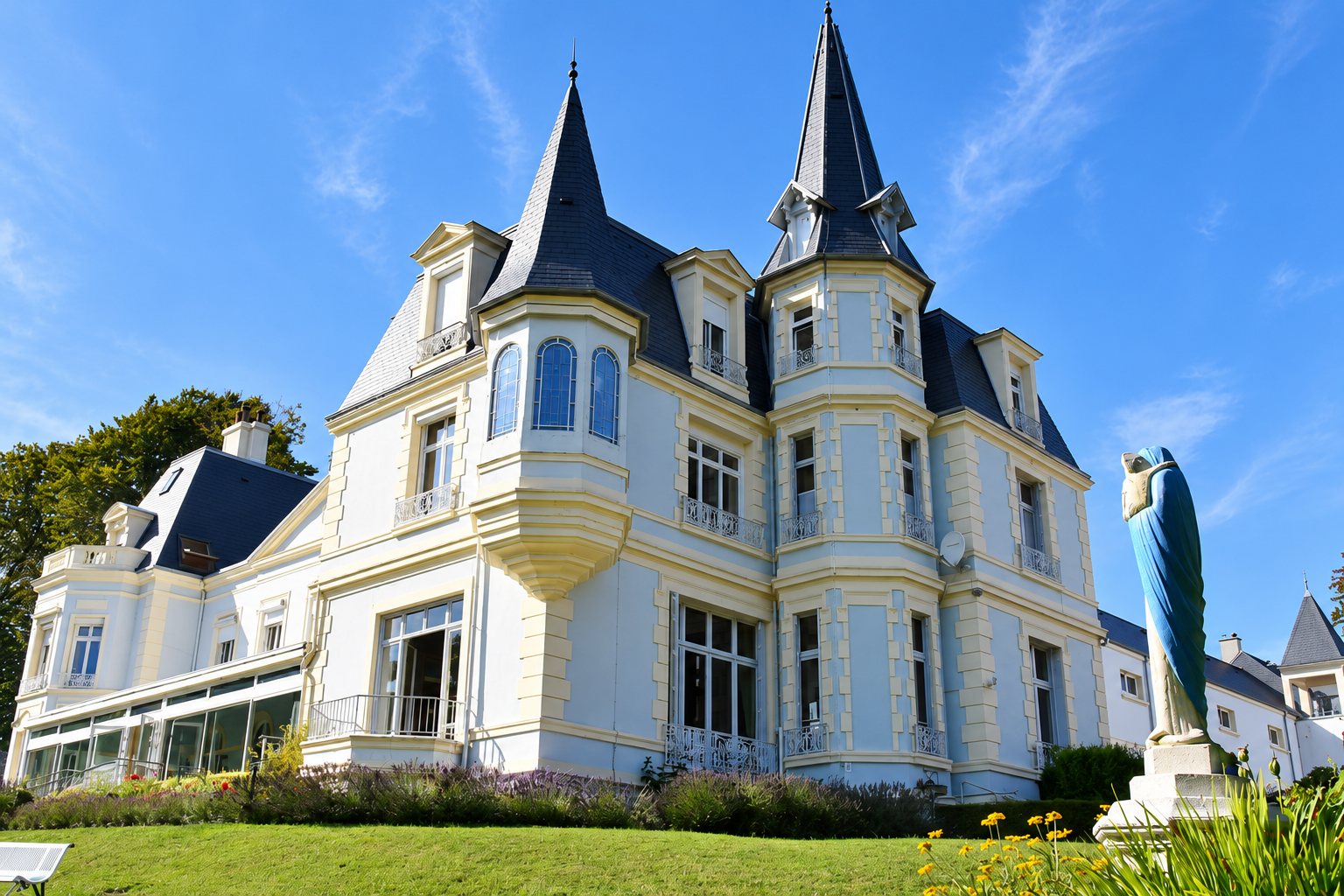 Façade d'un château de style néo-Renaissance aux murs bleu clair et aux tourelles couvertes d'ardoise, avec une statue bleue au premier plan et une pelouse fleurie sous un ciel bleu.