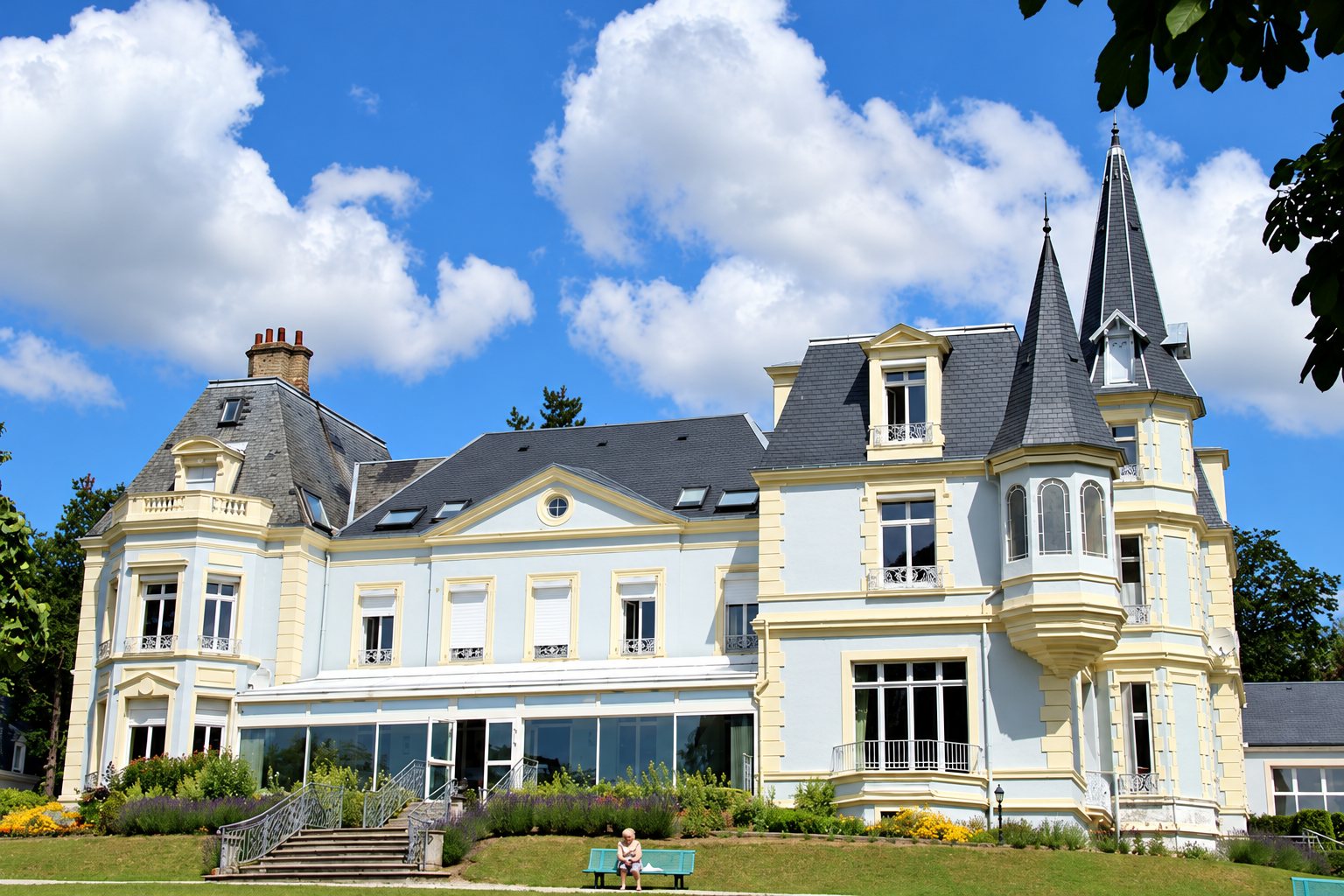 Façade d'un grand château de style néo-classique aux murs bleu pâle et aux toits d'ardoise, avec tourelles, escalier et jardin fleuri, sous un ciel bleu nuageux.