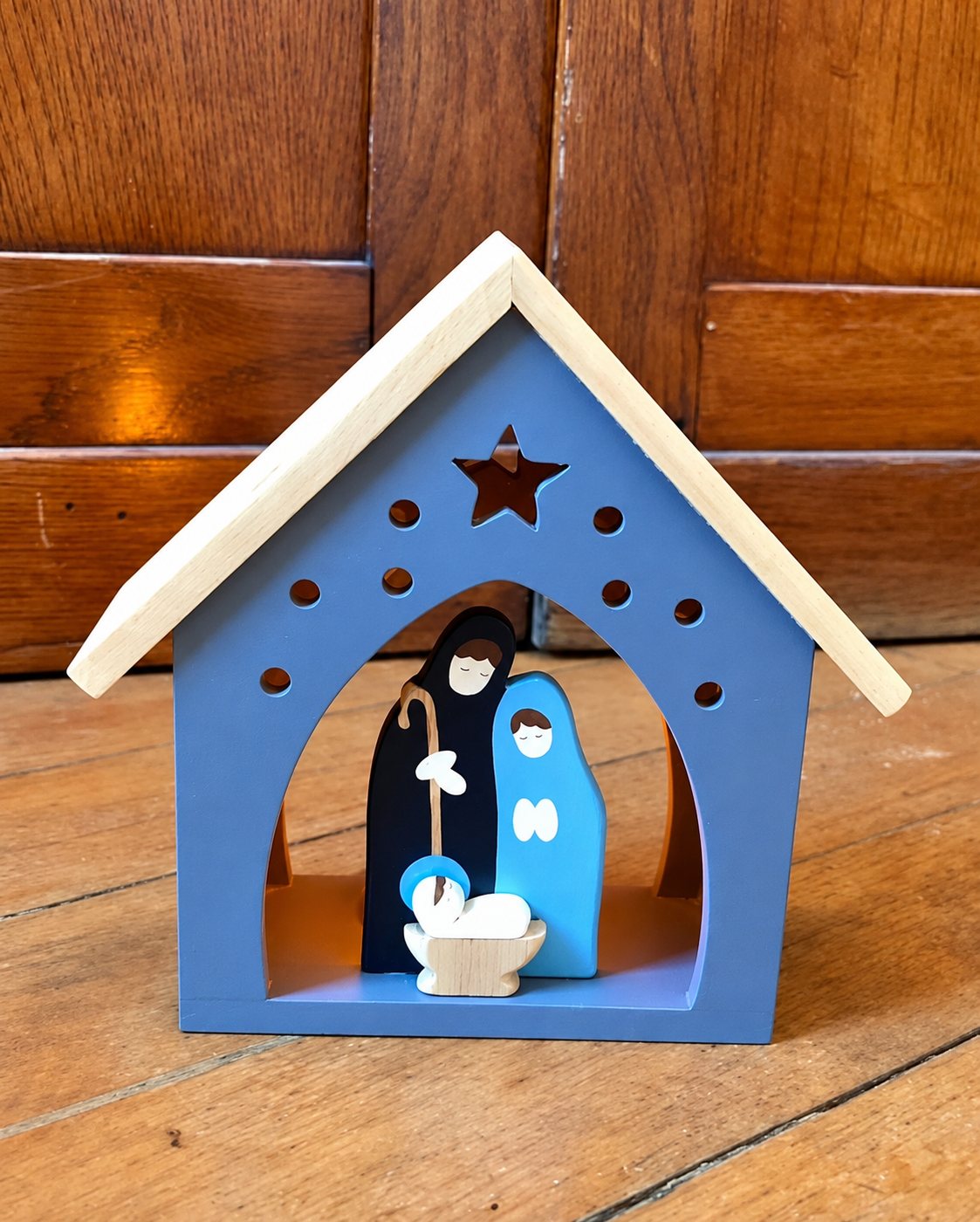 Petite crèche de Noël en bois peint en bleu-gris, avec une étoile découpée sur la façade, abritant Marie, Joseph et l'enfant Jésus dans sa mangeoire, posée sur un parquet devant une boiserie.
