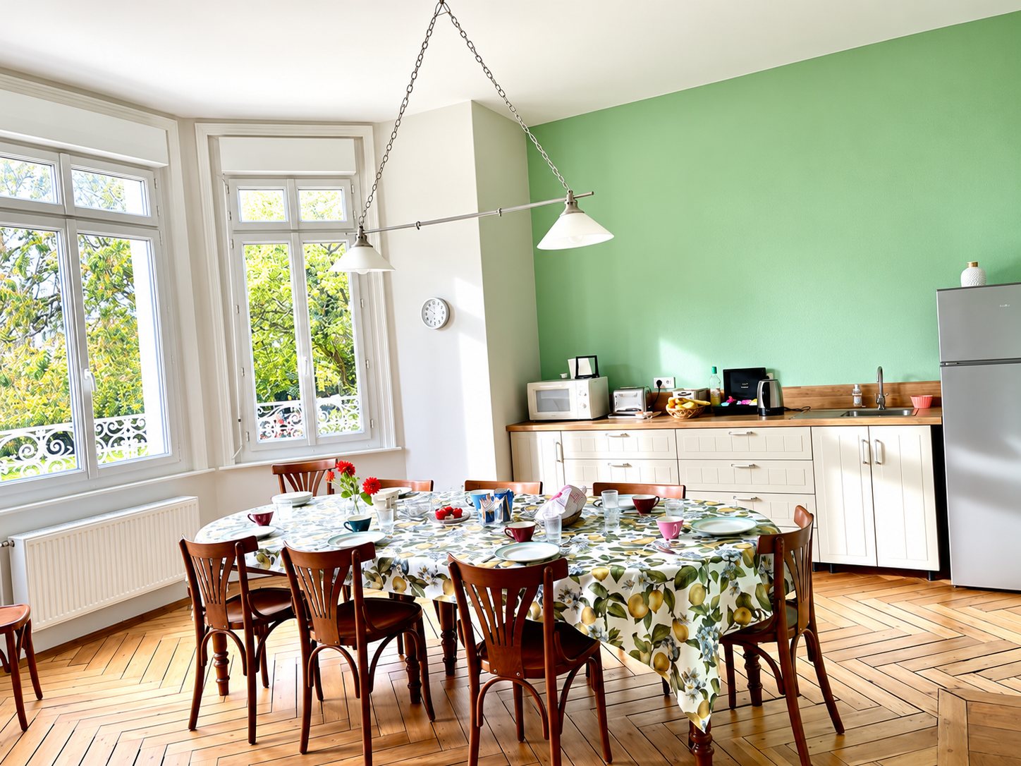 Une cuisine aux murs verts avec parquet en point de Hongrie, grandes fenêtres et une grande table ronde dressée avec une nappe à motif de citrons.