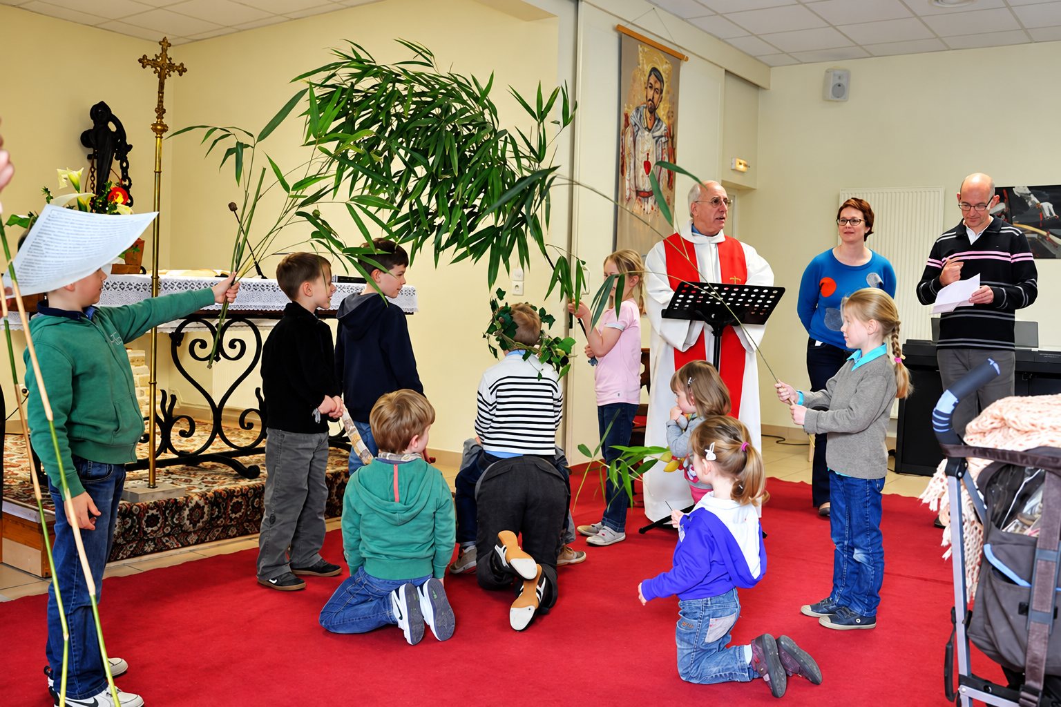 Des enfants tenant de longues branches vertes se rassemblent autour d'un prêtre en vêtements liturgiques rouge et blanc, dans une chapelle au sol recouvert de moquette rouge.
