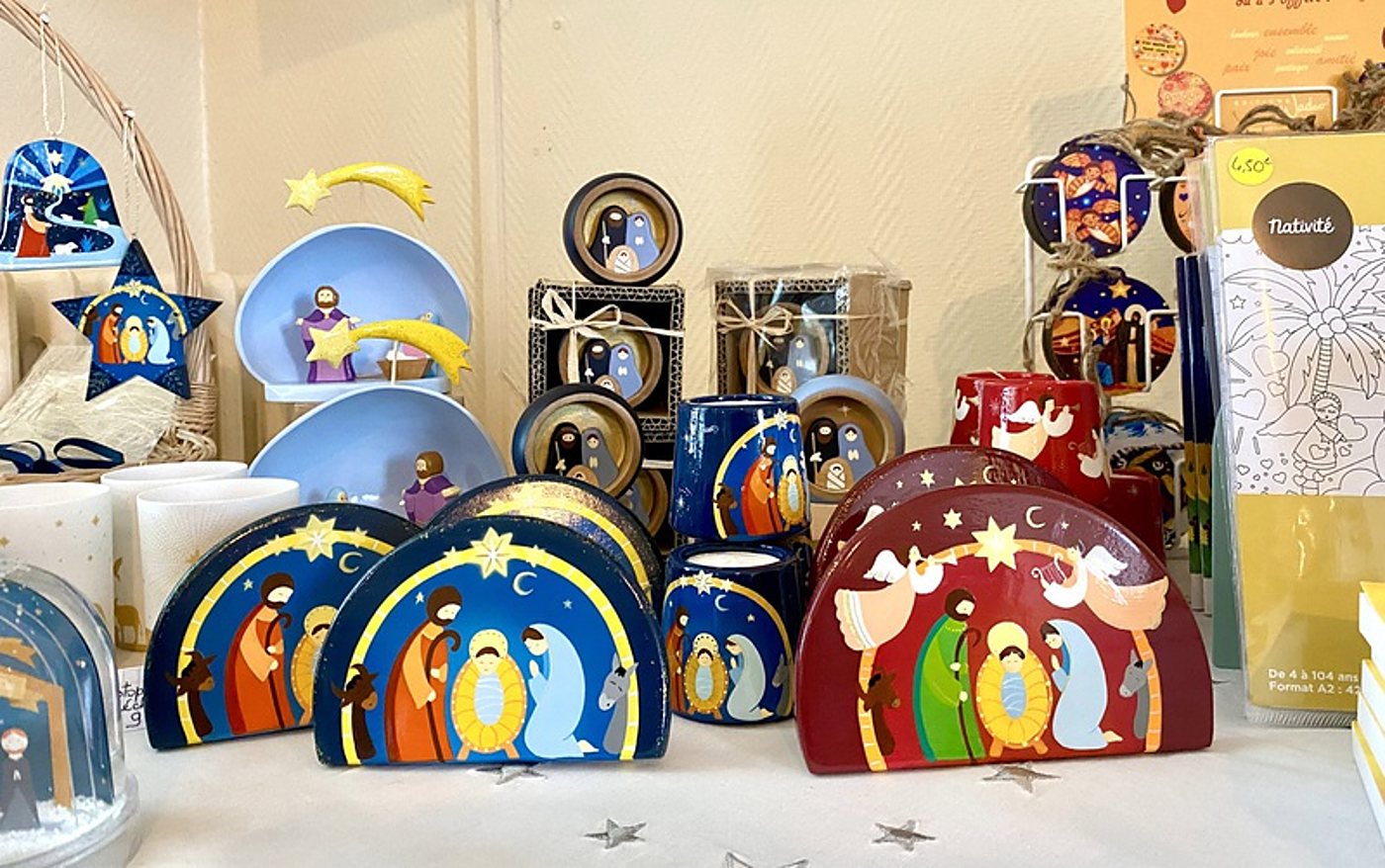 Sur une table décorée d'étoiles argentées, des objets de Noël colorés ornés de scènes de nativité, dont des bougeoirs en céramique bleus et rouges, sont présentés avec des étoiles filantes et des cartes.
