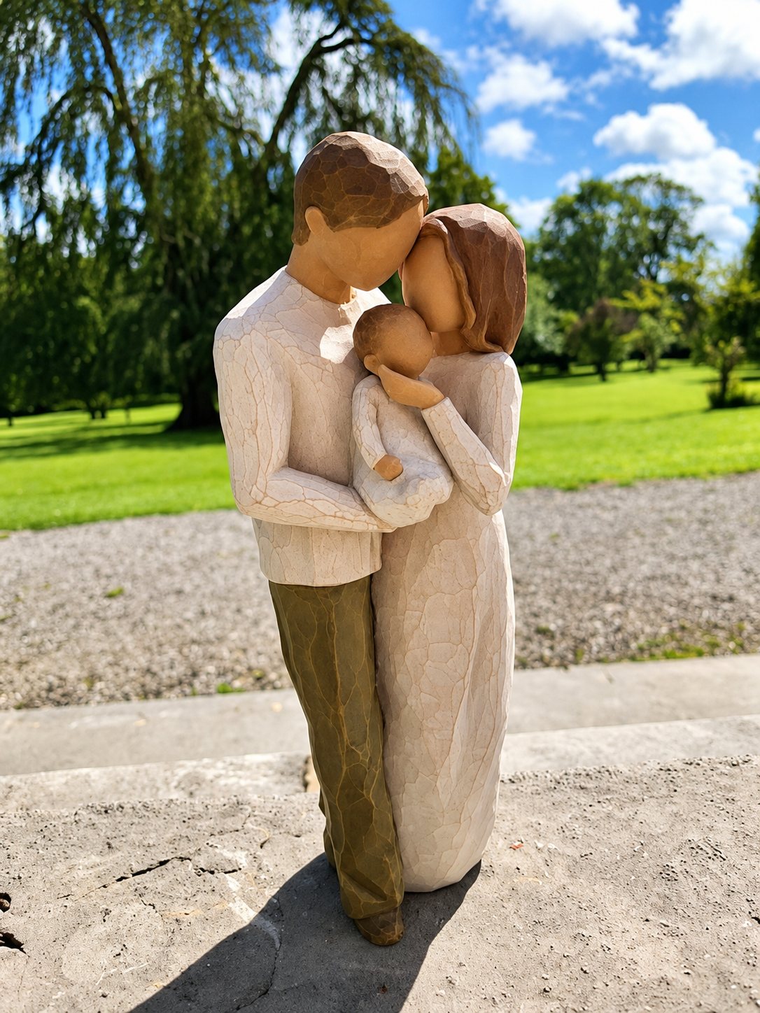 Une figurine sculptée représentant un homme et une femme penchés l'un vers l'autre et tenant tendrement un bébé, posée sur une pierre devant un parc verdoyant.
