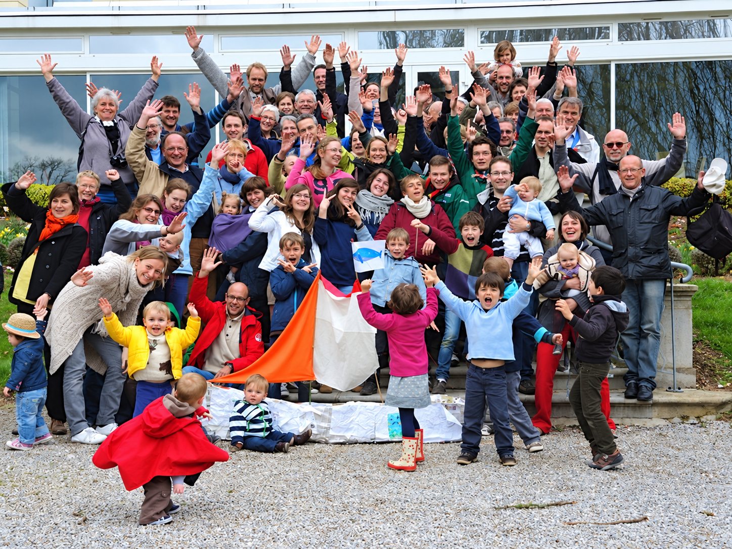 Un grand groupe d'adultes et d'enfants lève les bras en riant devant un bâtiment vitré, avec un drapeau orange et blanc au premier plan.