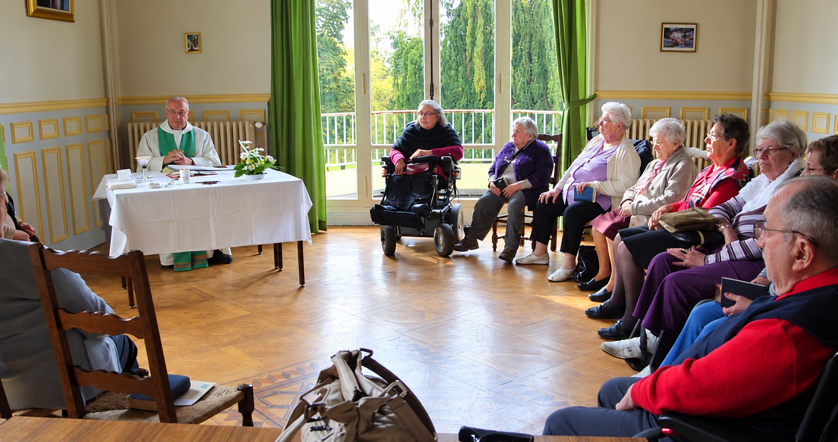Un prêtre en vêtements liturgiques verts célèbre une messe à une table drapée de blanc, devant des personnes âgées assises dans un salon lumineux aux grandes portes-fenêtres.
