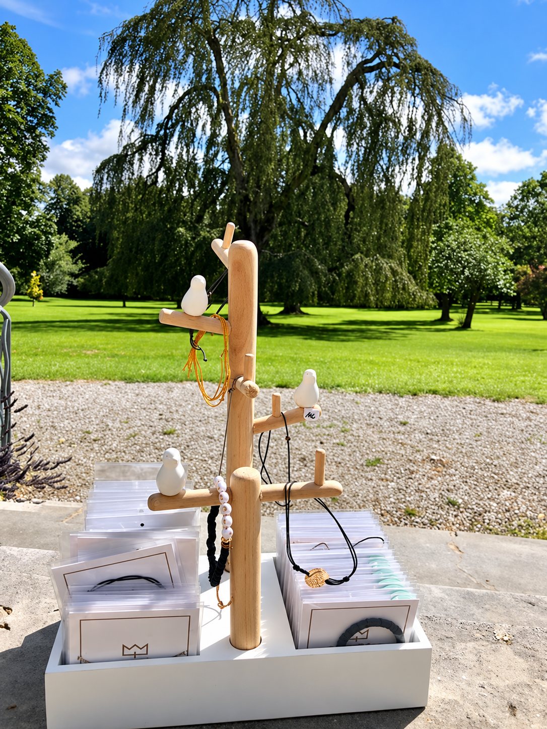 Un présentoir en bois en forme d'arbre, décoré de petits oiseaux blancs et de bijoux, posé sur une caisse blanche remplie de pochettes devant un grand parc verdoyant.