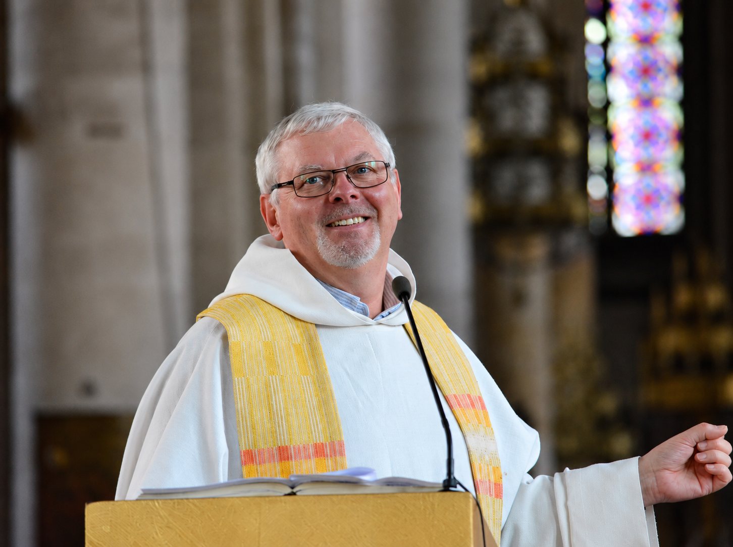 Un prêtre aux cheveux gris et à lunettes sourit derrière un pupitre doré, vêtu d'une aube blanche et d'une étole jaune, dans une église avec un vitrail en arrière-plan.