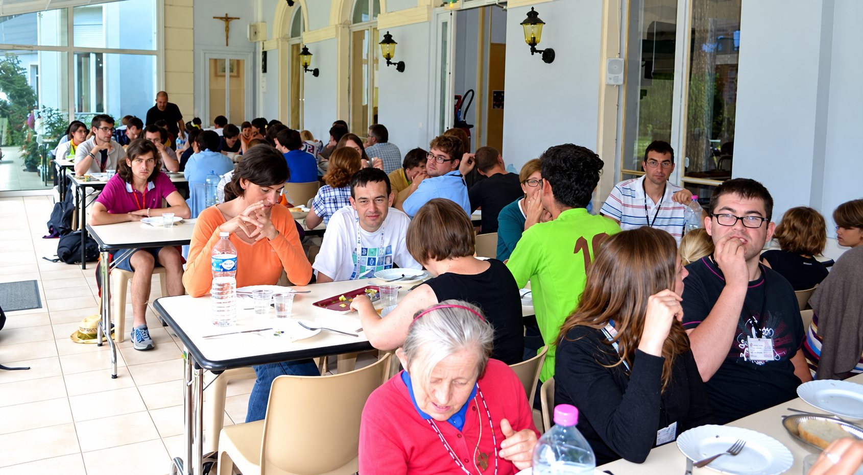 Des personnes de tous âges déjeunent ensemble à de longues tables dans une grande salle claire aux larges baies vitrées, avec un crucifix au mur du fond.