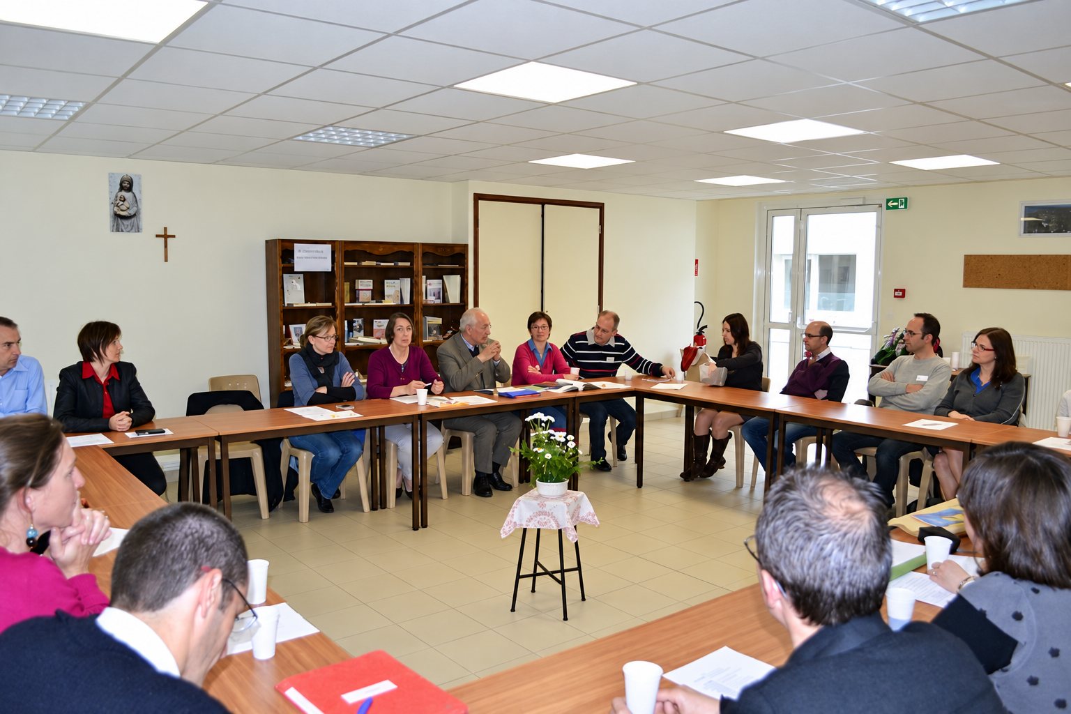 Une vingtaine de personnes sont assises autour de tables en carré dans une salle lumineuse, avec un bouquet de fleurs blanches au centre et une croix au mur.