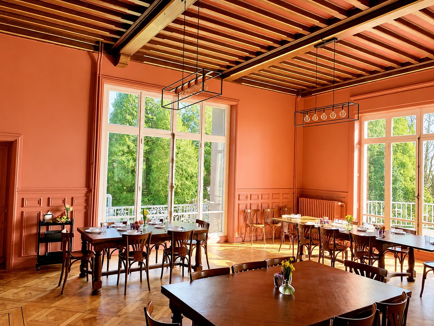 Une salle à manger aux murs orangés avec poutres apparentes, parquet et tables en bois dressées devant de grandes fenêtres donnant sur un parc.