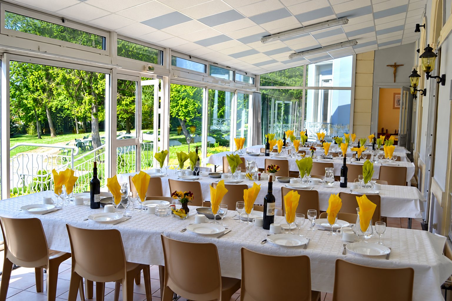 Une véranda lumineuse donnant sur un jardin, avec de longues tables couvertes de nappes blanches, de serviettes jaunes pliées, de verres et de bouteilles de vin.