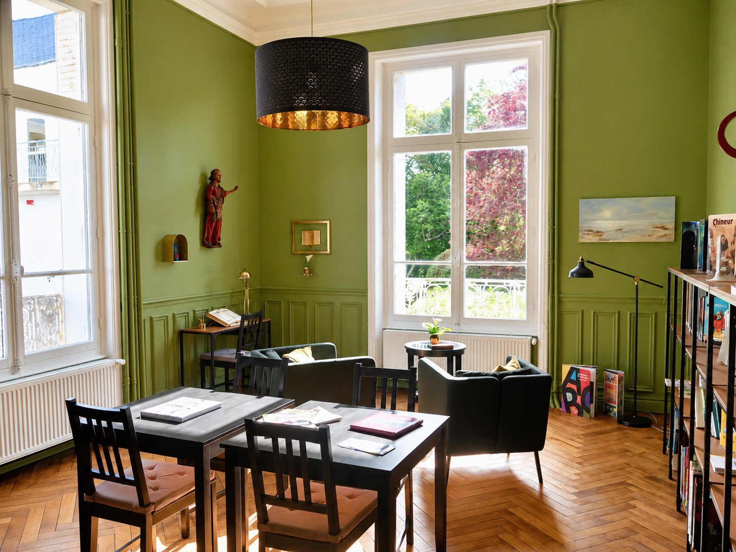 Une pièce lumineuse aux murs verts et au parquet en point de Hongrie, avec des tables en bois, des fauteuils, de grandes fenêtres et une bibliothèque remplie de livres.