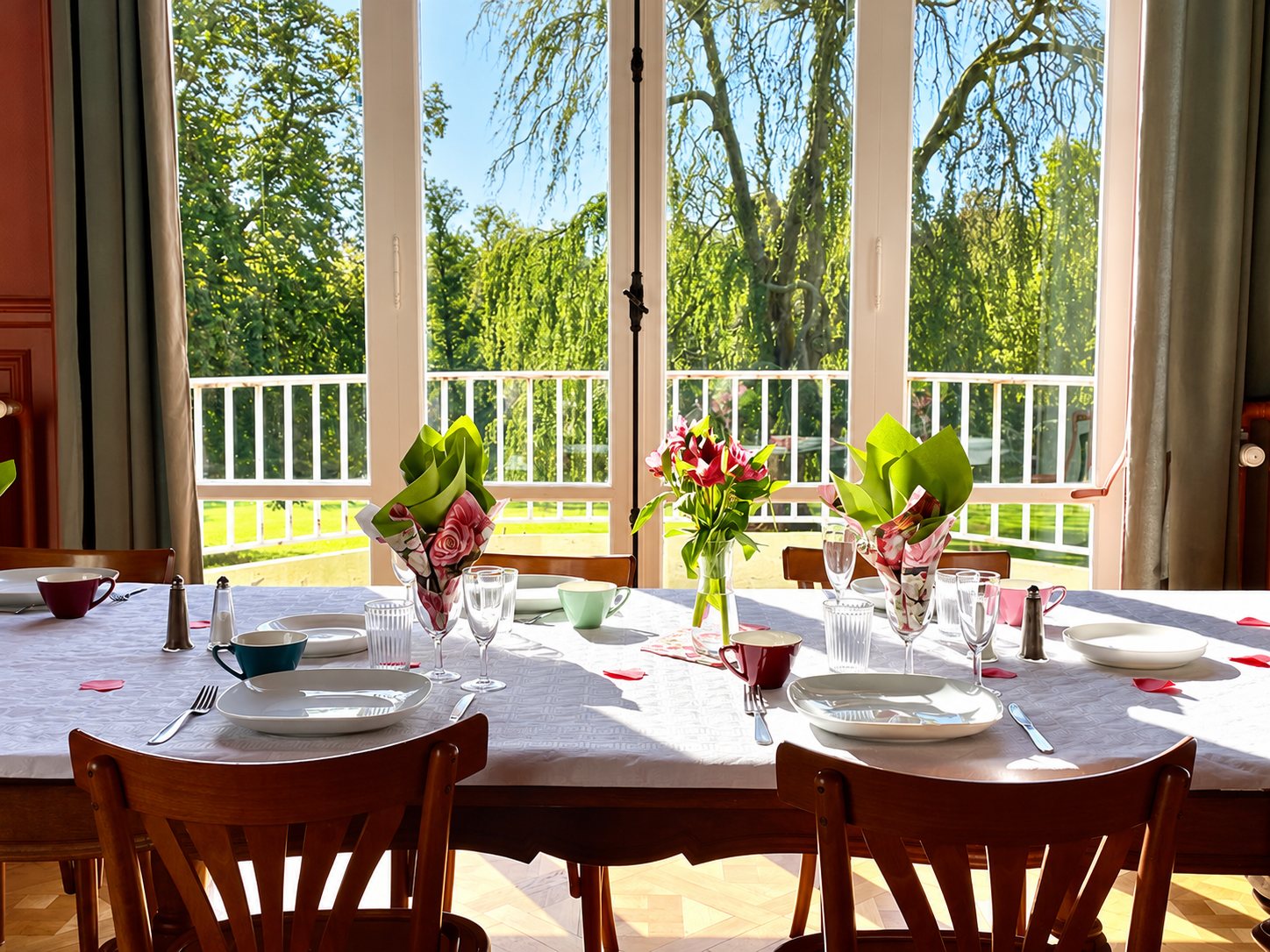 Une longue table dressée avec assiettes, tasses colorées, verres et petits bouquets de fleurs, face à de grandes fenêtres ouvrant sur un parc verdoyant et un balcon blanc.