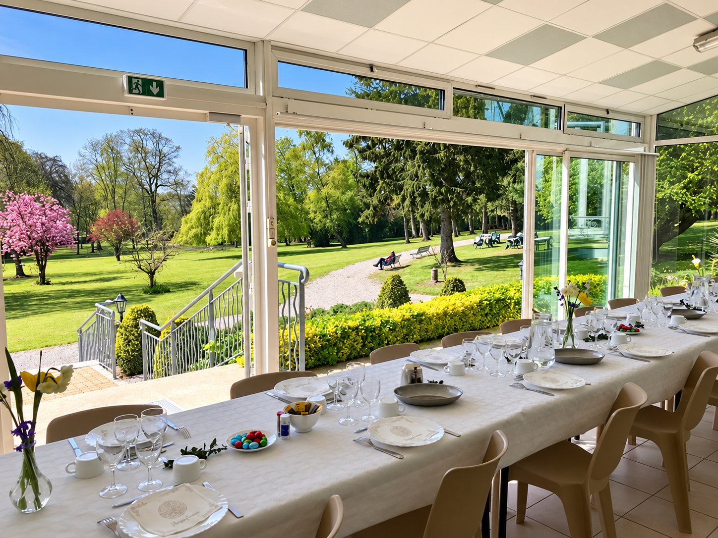 Une longue table blanche dressée avec verres, assiettes et fleurs dans une véranda lumineuse ouverte sur un grand parc arboré au printemps.